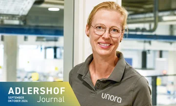 A smiling woman wearing glasses and a grey polo shirt with the word ‘unora’ on it, against a light-coloured, industrial background