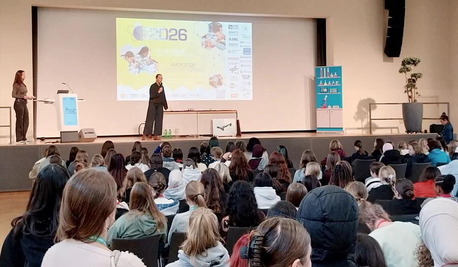 Vortragender steht auf Bühne vor großem Publikum in einem Saal mit Leinwand und Präsentation