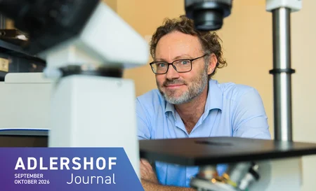 A man wearing glasses and a shirt is sitting in a laboratory, smiling, in front of a microscope