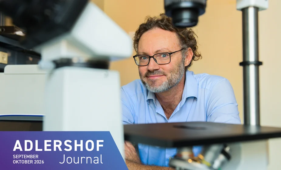 A man wearing glasses and a shirt is sitting in a laboratory, smiling, in front of a microscope
