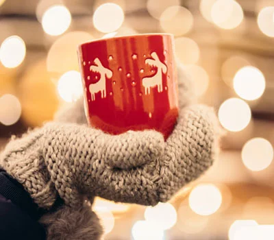 Hand in grauem Strickhandschuh hält rote Tasse mit weißen Rentiermotiven vor unscharfem Hintergrund mit Lichterketten