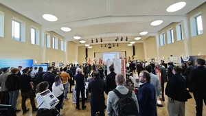 Großer Saal mit vielen stehenden Personen, die einer Präsentation auf einer Bühne mit Leinwand folgen