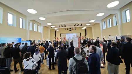 Großer Saal mit vielen stehenden Personen, die einer Präsentation auf einer Bühne mit Leinwand folgen