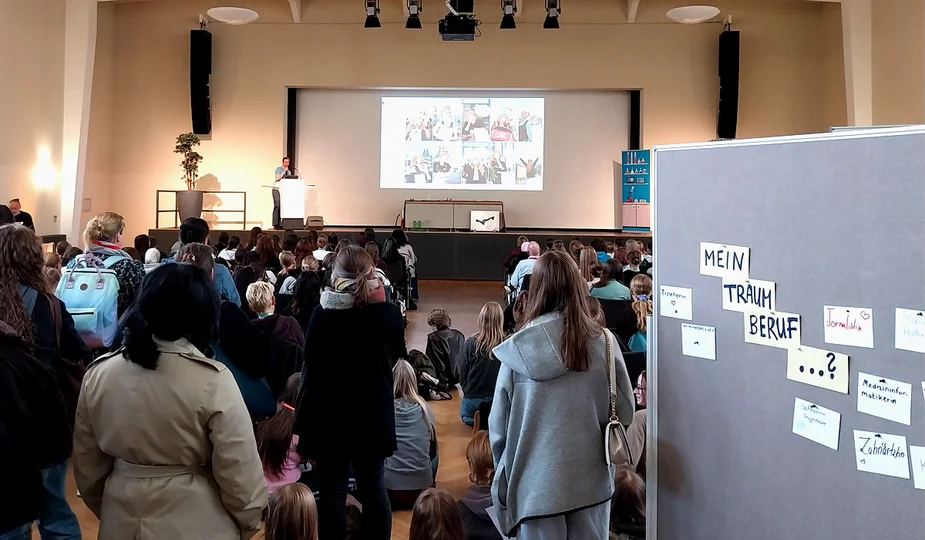 Menschenmenge in einem Saal hört einem Redner auf einer Bühne zu, rechts eine Pinnwand mit Zetteln und der Überschrift 'MEIN TRAUM BERUF ...?'