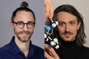 Portrait of two men side by side: the one on the left wears glasses and has a bun, while the one on the right has long hair and is wearing a black turtleneck sweater; he is holding a molecular model