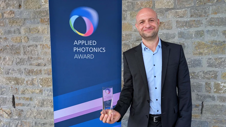 Marc Christ mit dem gläsernen Preis vor einem Banner mit der Aufschrift "Applied Photonics Award"